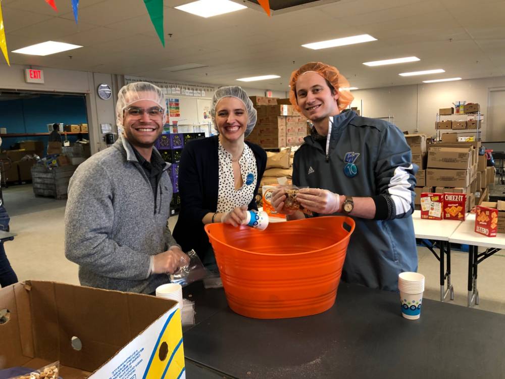 Three alumni smile for a photo while sorting food at Kids' Food Basket